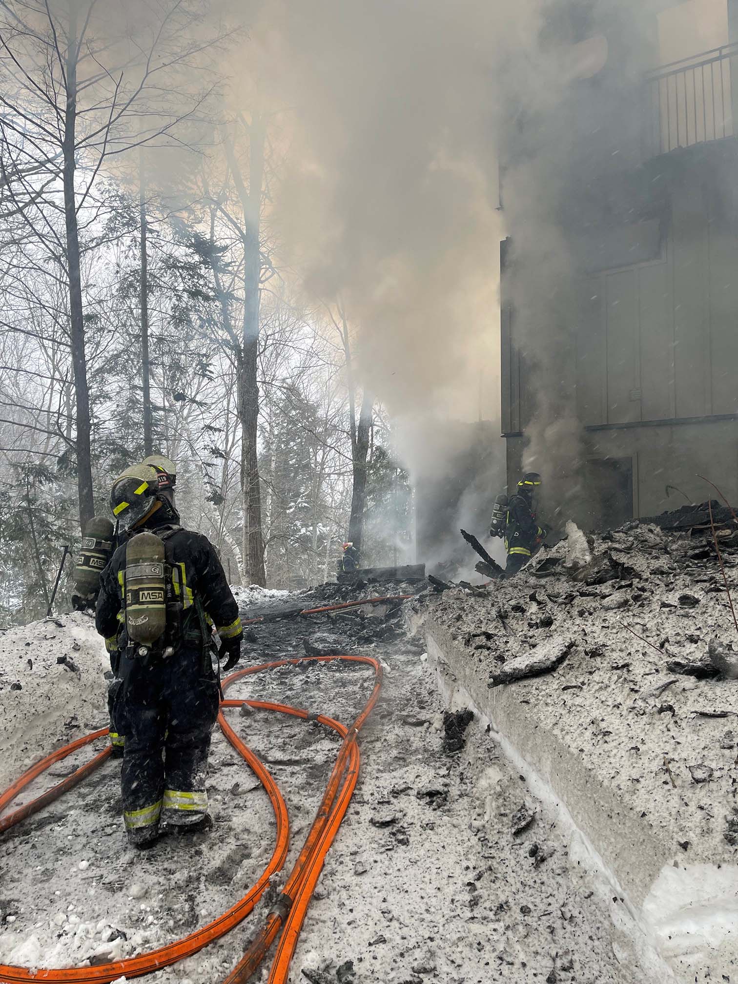 Firefighters extinguish a structure fire with heavy smoke and charred debris in the snow.